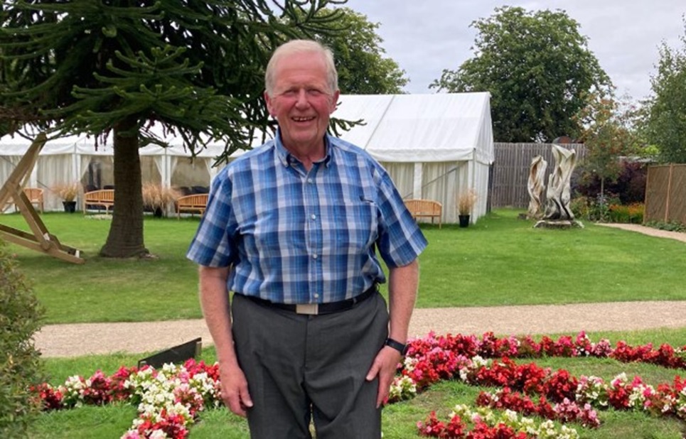 A photograph of Peter Dyde, a participant taking part in a BHF clinical trial .Peter is standing in a garden smiling at the camera. He is wearing a blue checked shirt with grey trousers. Behind him we can see a tree, a marquee and benches.