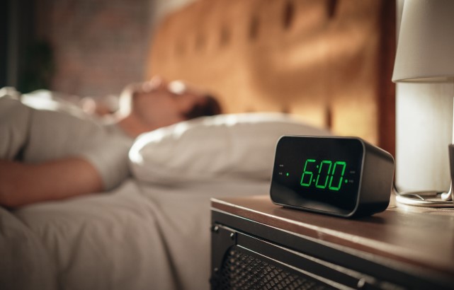 In the foreground of the photo there is a digital alarm clock sat on a table, showing the time 6:00. The table is next to a bed, and in the background of the photo there is a person asleep lying on their back under the covers with their head on a pillow.