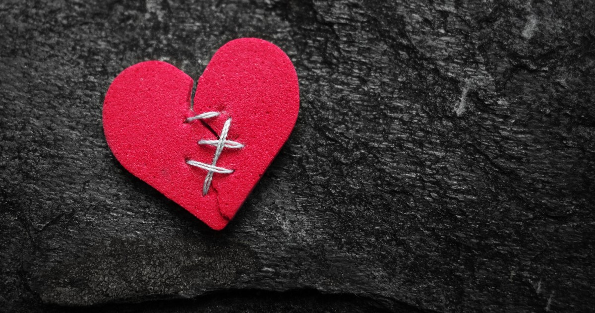 A red heart against a black background. The heart has been torn in half down the middle and there are a few stitches joining it back together.