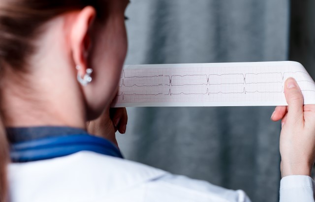 A photograph looking over the shoulder of a woman wearing a white lab coat as she reads a print out of an ECG trace