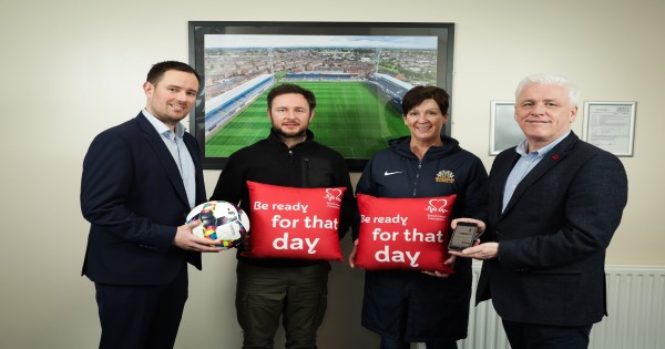 3 men and 1 woman standing against picture of Mourneview Park football pitch. 1 man holding football, 1 man holding phone and 3rd man and woman holding red cushions saying 'Be ready for that day'
