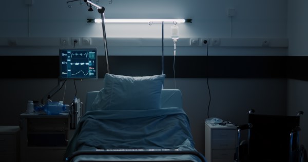 A photograph of an empty hospital bed in a dimly lit ward. There are two pillows sat on top of the bed with blue bedding covering the rest of the bed. There is a wheelchair sat empty on one side of the bed and a screen to monitor a patients' vital signs on the other above a table.