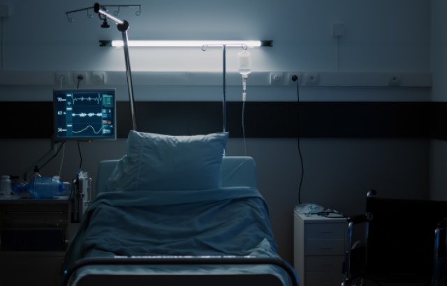 A photograph of an empty hospital bed in a dimly lit ward. There are two pillows sat on top of the bed with blue bedding covering the rest of the bed. There is a wheelchair sat empty on one side of the bed and a screen to monitor a patients' vital signs on the other above a table.