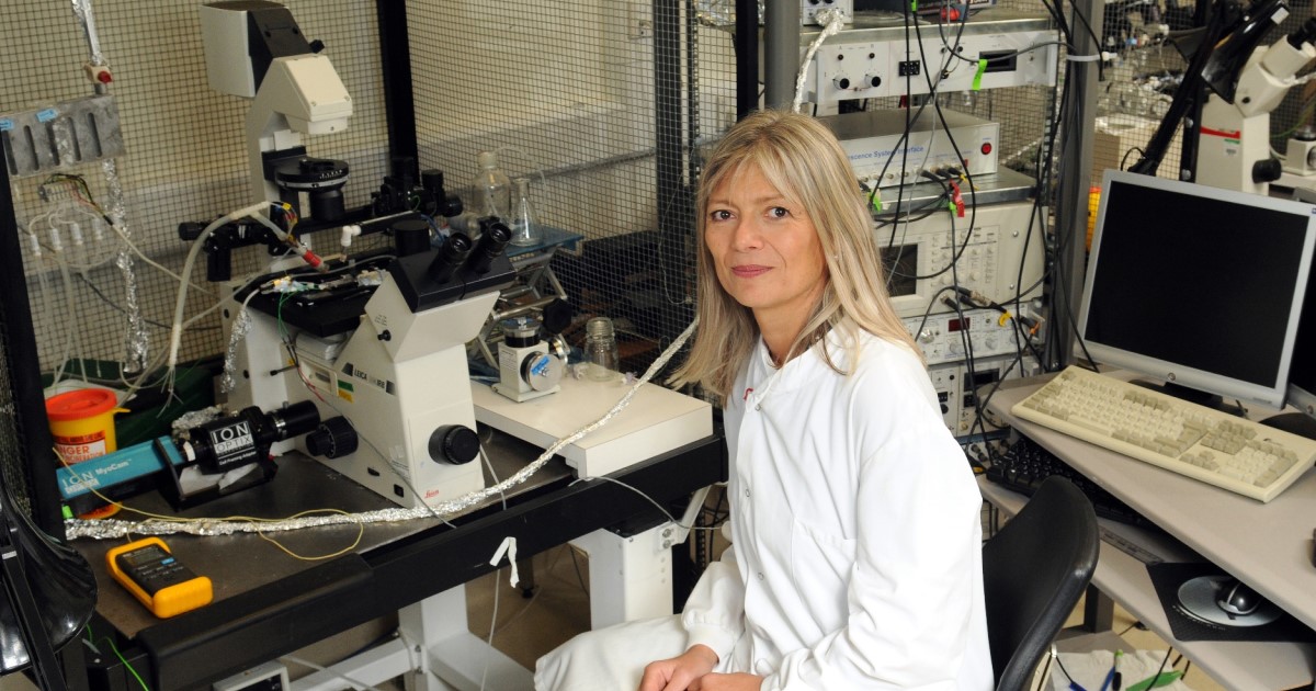 Photo of Professor Barbara Casadei sat in front of a large microscope. She is wearing a white lab coat and smiling at the camera. Behind her we can see a computer screen and keyboard, more microscopes and various equipment.