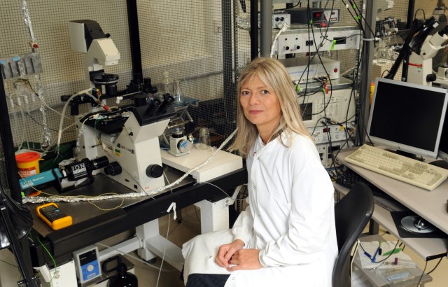 Photo of Professor Barbara Casadei sat in front of a large microscope. She is wearing a white lab coat and smiling at the camera. Behind her we can see a computer screen and keyboard, more microscopes and various equipment.