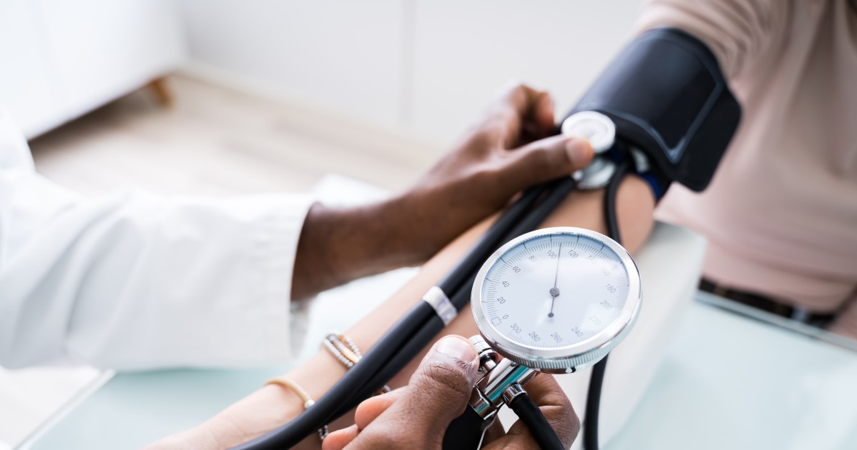 A photograph of someone wearing a white coat taking someone's blood pressure. They have a blood pressure monitor around their arm and the doctor is holding a stethoscope against the crease of their elbow and holding a pressure gauge in their other hand.