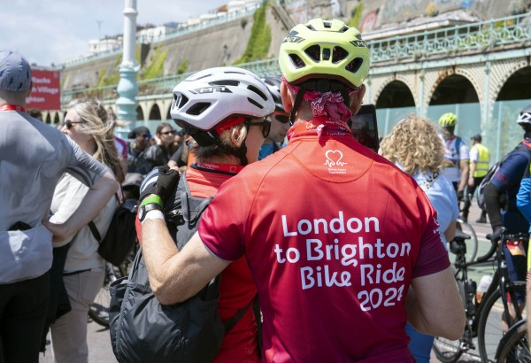 A male cyclist wearing a yellow-green helmet has his back turned to the camera. He's wearing a red jersey with white writing that says 'London to Brighton Bike Ride 2024;. He has his arm around another female cyclist wearing a silver helmet and a black backpack on her back. They are standing in a crowd of people near the arches on Brighton Beach