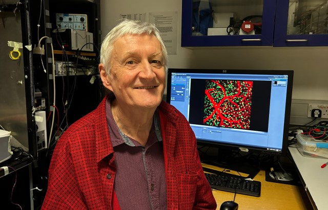 A man in a red shirt stands in a lab, in front of a screen showing a scientific image of fluorescent blood vessels