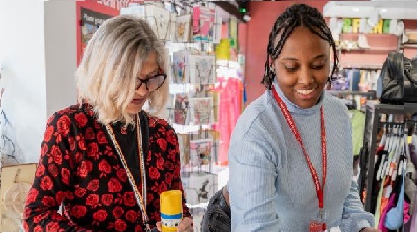 A white blond haired woman in a red and black spotty dress stands next to a young black woman with tied back braided hair. She is wearing a red BHF lanyard and wearing a grey jumper. They are in a BHF shop and both are looking down at something off camera.