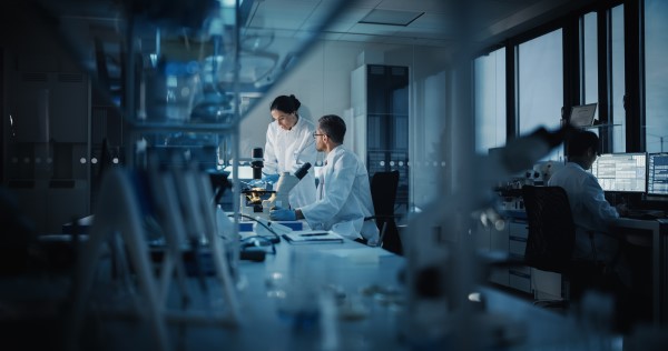 Photograph of a laboratory. In the foreground we can see a white lab bench with a microscope and various boxes. In the background we can two scientists, a man and a woman, both wearing white lab coats standing in front of another microscope having a conversation. On the right hand side of the image we can see another scientist with their back to the camera sitting in front of a computer with two monitors displaying various windows of text.