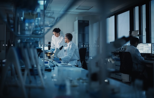 Photograph of a laboratory. In the foreground we can see a white lab bench with a microscope and various boxes. In the background we can two scientists, a man and a woman, both wearing white lab coats standing in front of another microscope having a conversation. On the right hand side of the image we can see another scientist with their back to the camera sitting in front of a computer with two monitors displaying various windows of text.