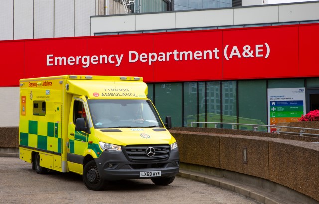 A yellow ambulance on a ramp in front of a red sign on a grey concrete building that reads: Emergency department (A&E)