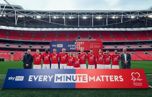 Photograph of the Sky Bet and BHF 'Re-Starting 11’ of football and community heroes including Graeme Souness, Glenn Hoddle, Tom Lockyer, Megan Tinsley, David Ginola and Fabrice Muamba. They are stood in Wembley Stadium, behind a sign that says 'Every Minute Matters'