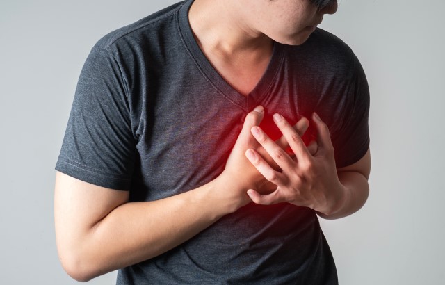 A man wearing a dark grey t-shirt clutches his chest. There is a red glow added to the area above his hands/heart