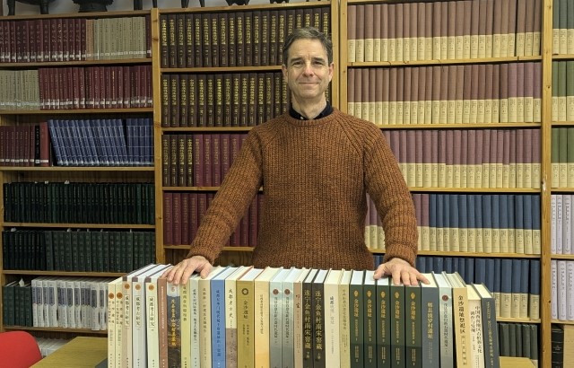 Trial participant John Moffett posses with a selection of books in front of a large bookcase 