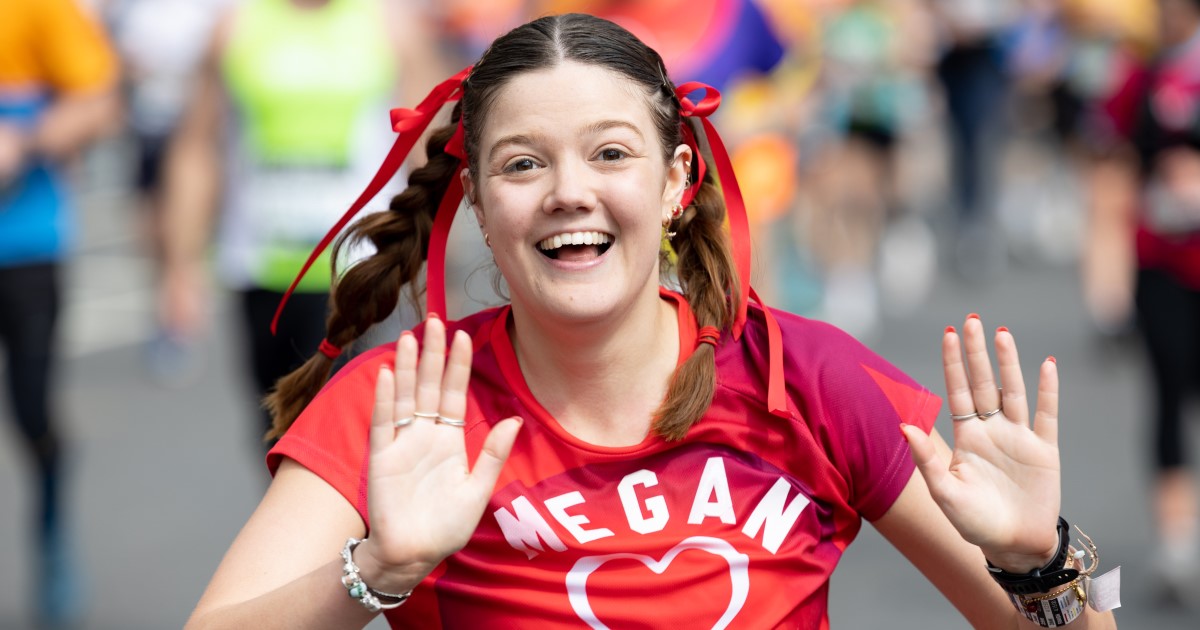 A woman running the London Marathon waves with both hands at the camera. She has a big smile on her face. Her hair is in two plaits with red ribbons. She is wearing a red British Heart Foundation runner top with her name Megan on it