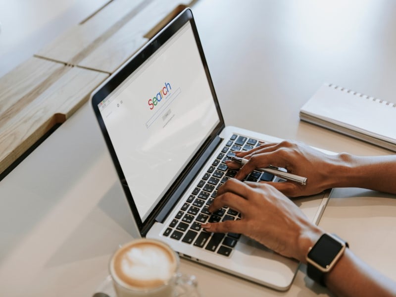 A par of disembodied hands typing at a sleek laptop, a Macbook Pro, on a pale desk. The left arm has an Apple Watch on the wrist. There is a coffee cup with foamed milk sitting to the left, partially out of view.