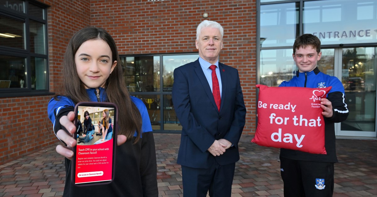 School students girl (left ) and boy (right) with Fearghal McKinney from British Heart Foundation in middle. Outside at front of school, girl holding mobile phone, boy holding cushion.