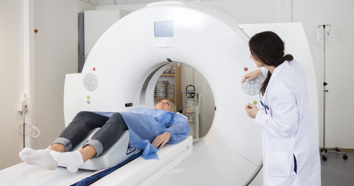 A doctor in a while coat looks at a patient as they lie on a the bed of a CT scanner