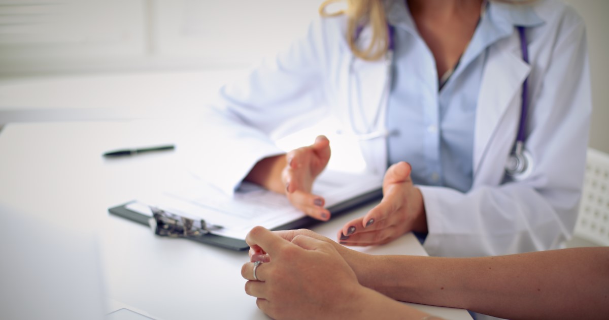 Photograph of a doctor in a white coat sitting at a table speaking to their patient. 