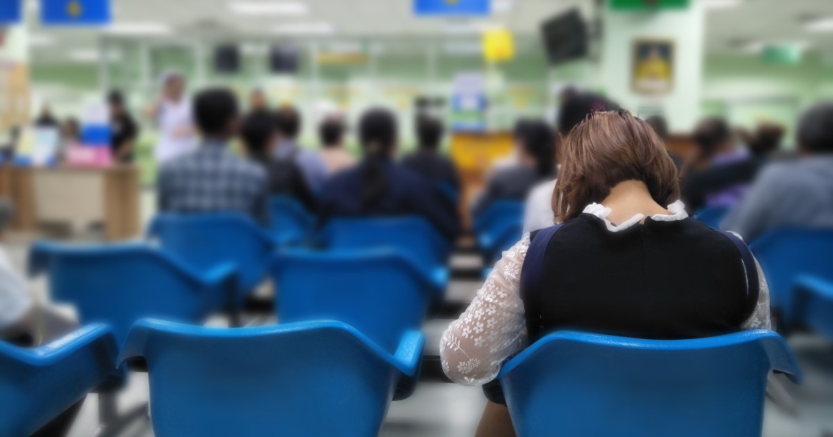 A woman sits with her back to us in a crowded hospital waiting room with her head down