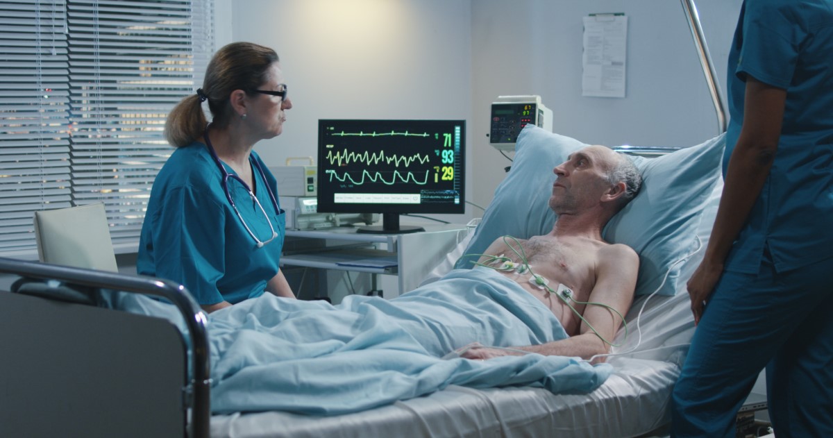 An elderly male patient with ECG cables on his body speaks to a female doctor 