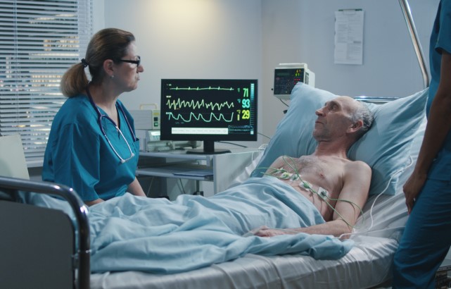 An elderly male patient with ECG cables on his body speaks to a female doctor 