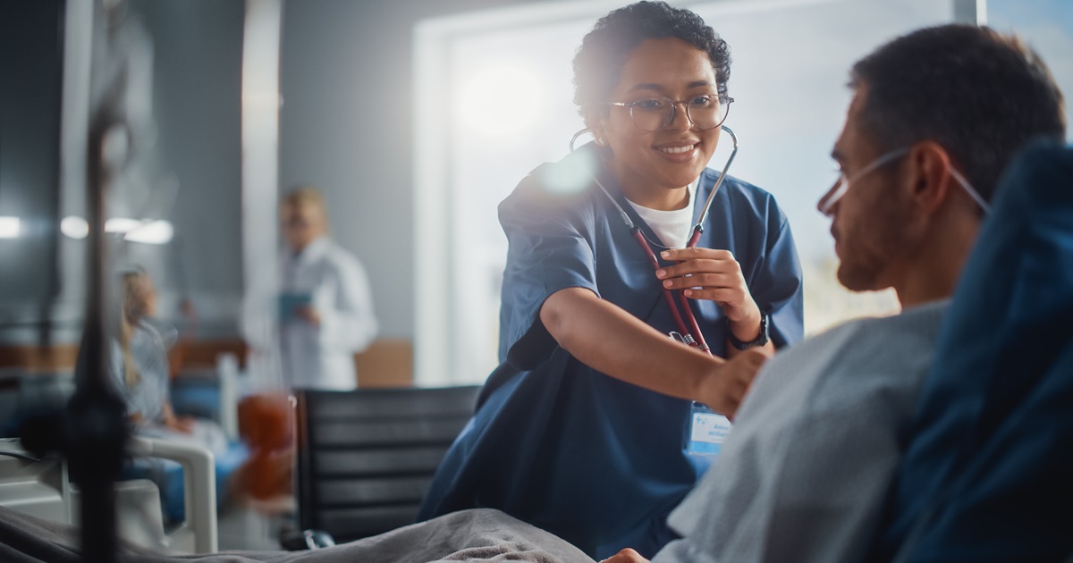 A doctor holds a stethoscope to a patient's chest. They are sitting in a hospital bed and the doctor is stood next to them.