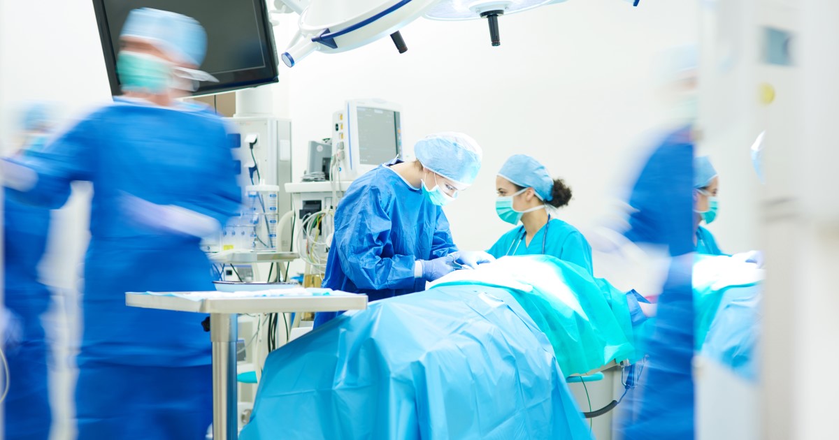 Photograph of an operating room, filled with doctors and health professionals in scrubs and wearing masks and gloves.