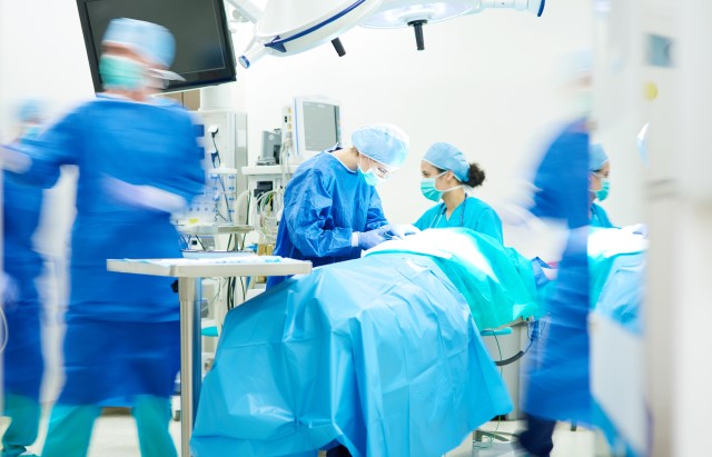 Photograph of an operating room, filled with doctors and health professionals in scrubs and wearing masks and gloves.
