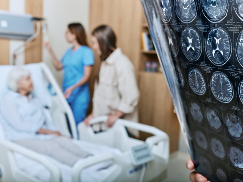 An elderly woman sits in a hospital bed. In the corner of the screen someone is reviewing an MRI brain scan images.