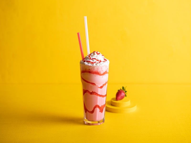 A strawberry milkshake with red strawberry sryup laced in a spiral around the tall glass it's in, with a red and white straw poking out the top. Just behind the glass is three slices of mango topped with a single strawberry. The backdrop is plain golden yellow.