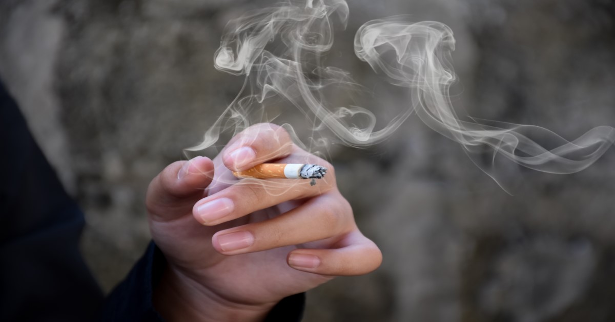 A disembodied hand is in the centre of frame, holding a smoking cigarette