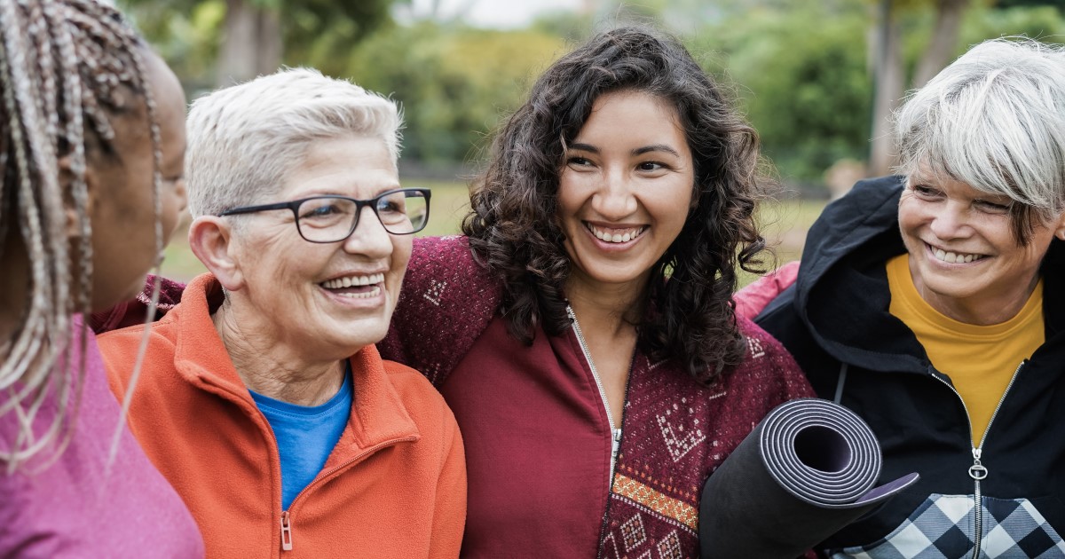 Four women in workout clothes stand in a row laughing and smiling with each other. A black women with long blonde and black braids has her face turned away from the camera, and is wearing a pink top. The second woman is White and has short white grey hair, wears black rimmeed glasses and is wearing an orange jacket zipped up with a blue top underneath. The third woman is mixed race and has shoulder length dark wavy hair and is smiling at the Black woman. She is wearing a dark red jacket and has a black yoga mat rolled up under her arm. The fourth woman has white grey hair with a sweeping side fringe tinged with dark grey, and is wearing a black cropped jacket which has a chequered black and white pouch over the stomach, it's zipped up with a yellow turtleneck top underneath. There are green leafed trees in the background.