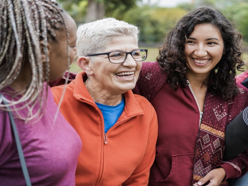 Three women in workout clothes stand in a row laughing and smiling with each other. A black women with long blonde and black braids has her face turned away from the camera, and is wearing a pink top. The second woman is White and has short white grey hair, wears black rimmeed glasses and is wearing an orange jacket zipped up with a blue top underneath. The third woman is mixed race and has shoulder length dark wavy hair and is smiling at the Black woman. She is wearing a dark red jacket and has a black yoga mat rolled up under her arm. There are green leafed trees in the background.