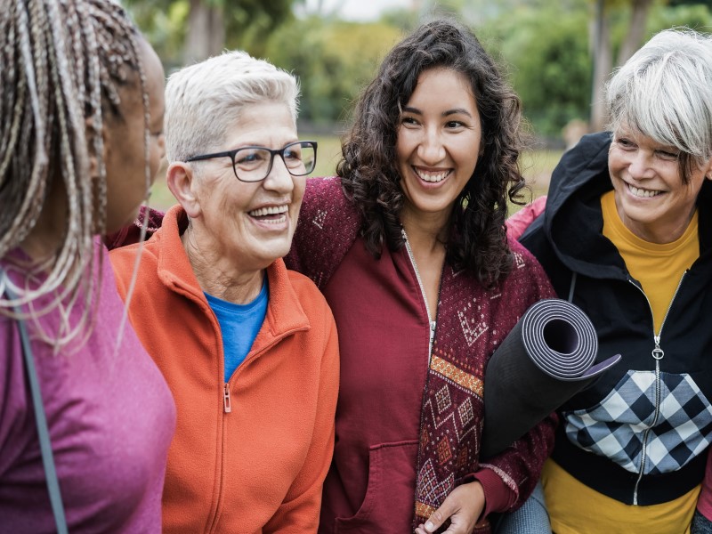 Four women in workout clothes stand in a row laughing and smiling with each other. A black women with long blonde and black braids has her face turned away from the camera, and is wearing a pink top. The second woman is White and has short white grey hair, wears black rimmeed glasses and is wearing an orange jacket zipped up with a blue top underneath. The third woman is mixed race and has shoulder length dark wavy hair and is smiling at the Black woman. She is wearing a dark red jacket and has a black yoga mat rolled up under her arm. The fourth woman has white grey hair with a sweeping side fringe tinged with dark grey, and is wearing a black cropped jacket which has a chequered black and white pouch over the stomach, it's zipped up with a yellow turtleneck top underneath. There are green leafed trees in the background.