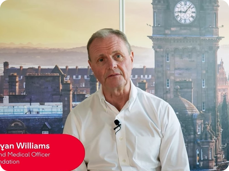 Professor Bryan Williams sits in front of artificial London backdrop with a microphone attached to his shirt.