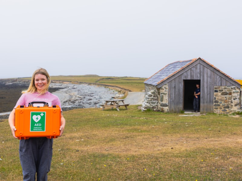A young blonde woman stands with an orange case containing a defibrillator on a remote Welsh island with a shed in the background. In the doorframe of the shed leans a young man. 