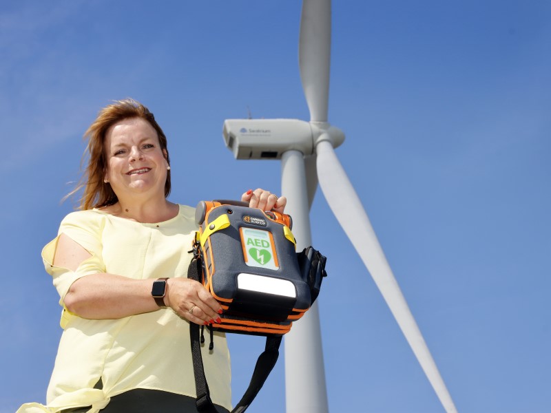 A white middle aged woman stands holding a defibrillator in a black soft case, there is a wind turbine in the backdrop