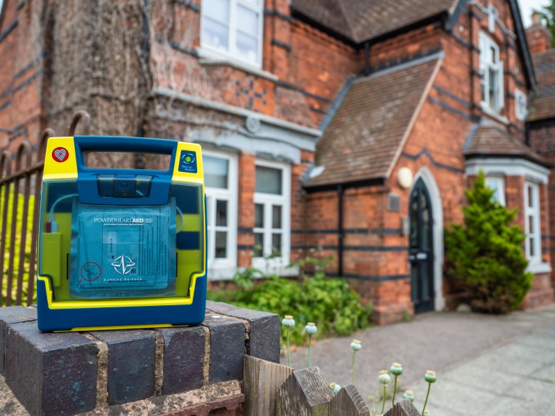 A blue-green defibrillator sits on a brick gatepost outside an old fashioned Victorian redbrick primary school