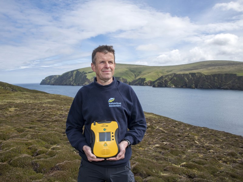 A white man stands with a yellow defibrillator in front of him, a beautiful Scottish loch is in the background overlookd by a lush green mountain