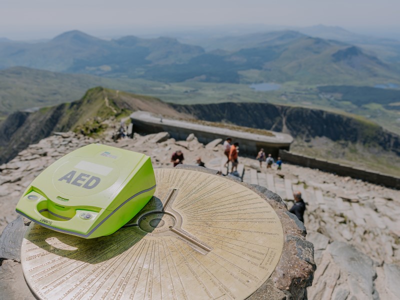 A light green defibrillator sits on a sundial at the top of Mount Snowdon, there is a stunning panoramic vista in the foreground