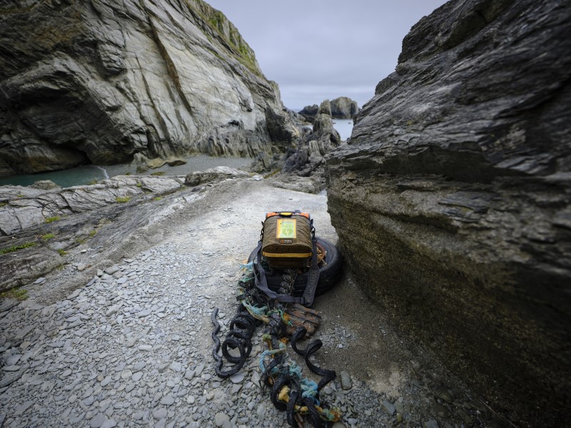 An orange defibrillator sits on an abandoned rubber buoy in an inlet by the sea, towered over by columns of rock