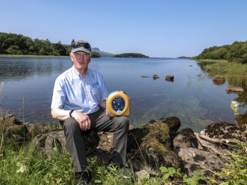 An elderly white man sits on a line of rocks by the shore of a lake, he is holding a yellow defibrillator in his left hand