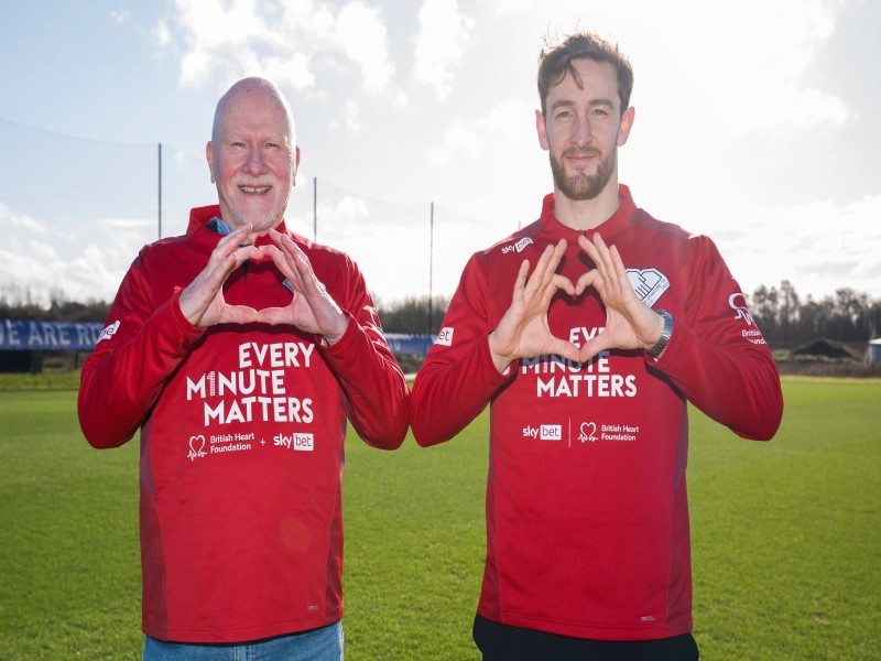 Image of cardiac arrest survivors Maurice Jones and Tom Lockyer standing on a football pitch making heart symbols with their hands 