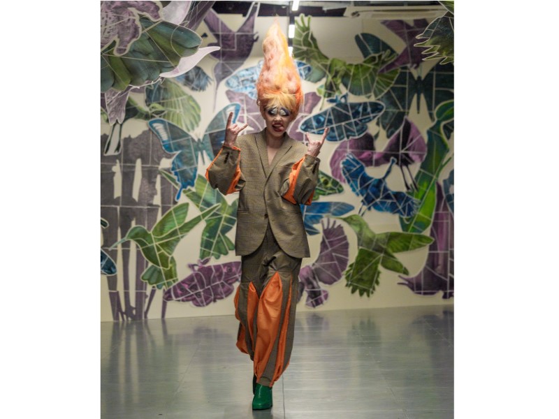 Model walking down the catwalk who has orange fair and is wearing an orange and green suit.