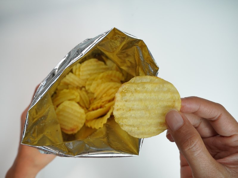 A unbranded open packet of crisps with a hand reaching into the packet to grab one ridged crisp, against a white backdrop