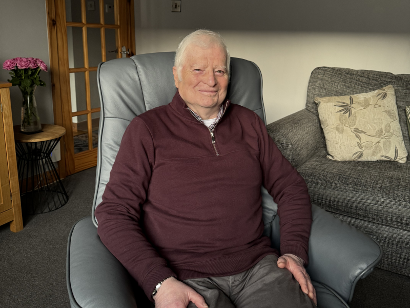 Photograph of John Macdonald, the first patient to receive a new gene therapy to prevent coronary artery bypass grafts failing. John is sitting in a chair smiling at the camera.