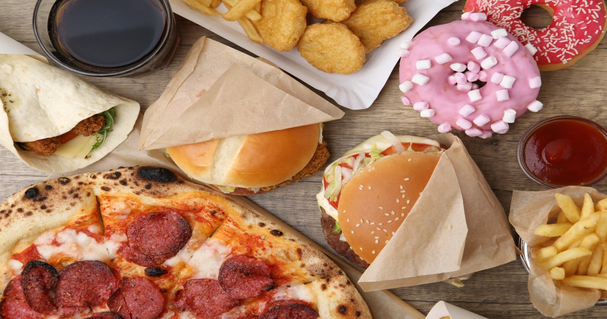 An array of junk food laid out on a table, from left to right in a circle is a pint glass of Coca Cola, some chicken nuggets, two pink glazed doughnuts, some ketchup in a little dish, a portion of fries, two burgers wrapped in brown napkins, and half a pepperoni pizza, and half a chicken wrap.