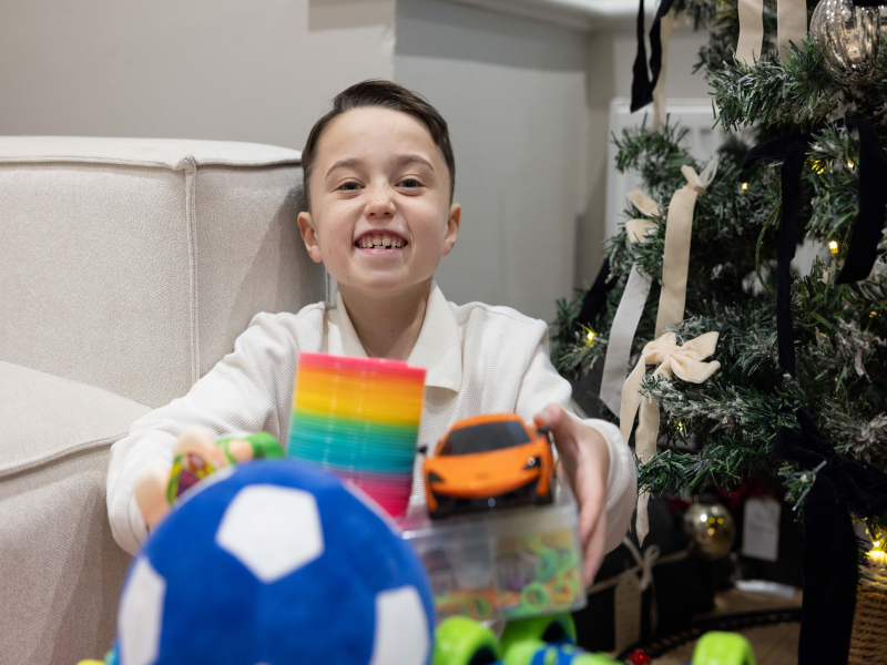 Eight year old young heart hero Raphie, sat next to a Christmas tree and holding presents, including an orange toy car and a blue football, towards the camera